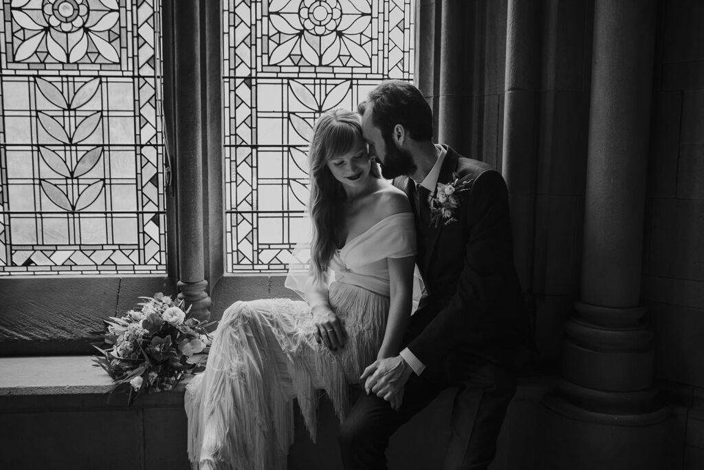 Black and white portrait of the bride and groom cuddling in front of the stained-glass window at the Whitworth Gallery in Manchester.